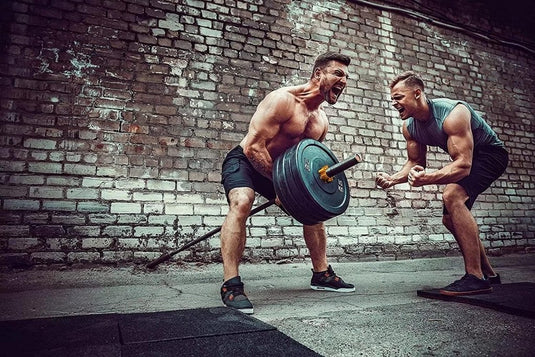 Een gespierde CrossFit-fanaat tilt een zwaar beladen halter op, terwijl een andere man hem energiek aanmoedigt. Het tafereel speelt zich af tegen een ruige bakstenen muur, waarbij alle intensiteit wordt vastgelegd door de betrouwbare grip van "Train professioneel met deze Olympische halterklemmen!.