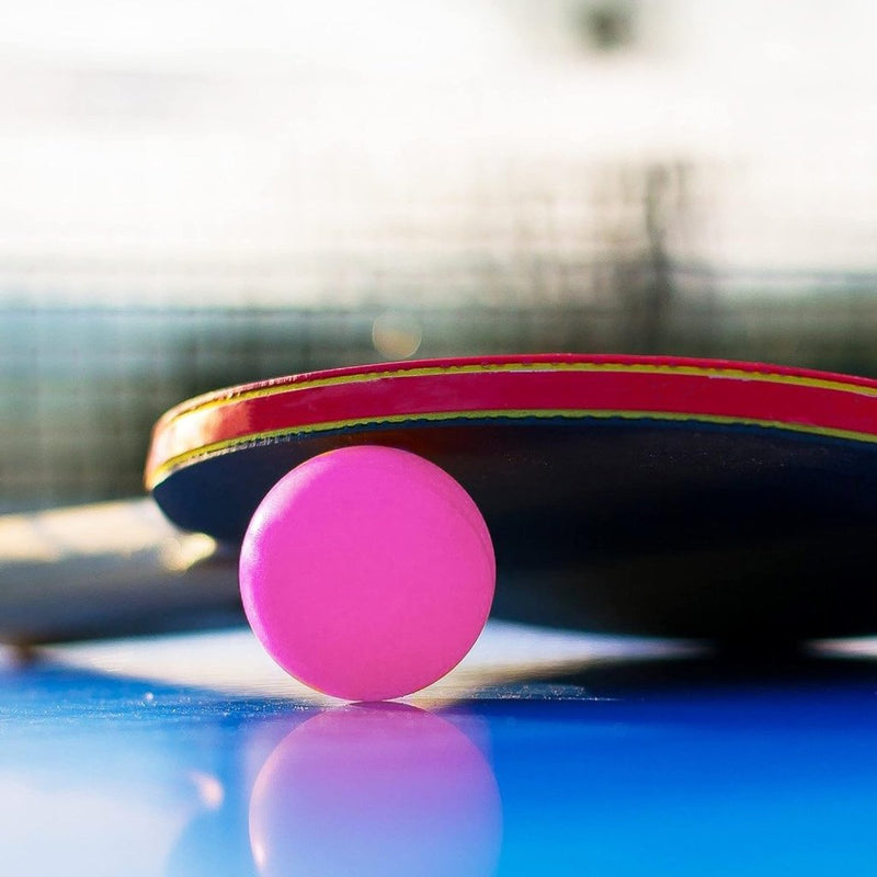 Laad afbeelding in Galerijviewer, Eén van de roze ballen uit &quot;De perfecte set van 24 tafeltennisballen – Voor pingpong, bierpong en meer!&quot; in standaardformaat 40 mm zit bij een peddel op een blauwe tafel, met daarachter een wazig net.
