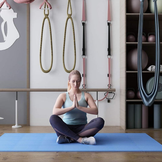 Een vrouw zit met gekruiste benen op een Transformeer je vloer met onze zachte interlockende puzzelmat, haar handen in gebedshouding, omringd door fitnessapparatuur en yoga-attributen die de serene sfeer versterken.