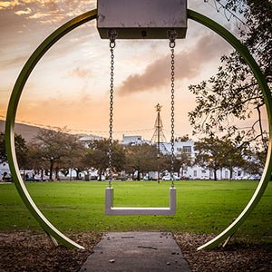 Load image into Gallery viewer, Een moderne, ronde schommel van roestvrij staal staat leeg in het park, omlijst door bomen, met een grasveld en stadsgebouwen op de achtergrond, onder een oranje lucht.
