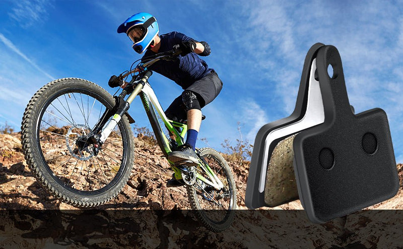 Load image into Gallery viewer, Een fietser in beschermende kleding navigeert vakkundig met een mountainbike van een rotsachtige helling, wat de duurzaamheid van Perfecte remblokken voor jouw mountainbike avonturen! laat zien! Een close-up toont de remblokken in actie tegen een achtergrond van een helderblauwe lucht.
