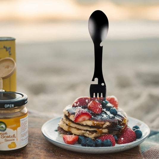 Een stapel pannenkoeken met aardbeien en bosbessen wordt elegant versierd met de "De ultieme multifunctionele campinglepel: Jouw beste vriend voor elk avontuur", geplaatst op een bord naast een pot butterscotchsaus, tegen een serene strandachtergrond.