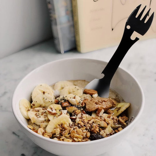Een kom havermout belegd met gesneden bananen, muesli, gehakte noten en zaden bevat de De ultieme multifunctionele campinglepel: Jouw beste vriend voor elk avontuur erin. Op de achtergrond zijn boeken zichtbaar.