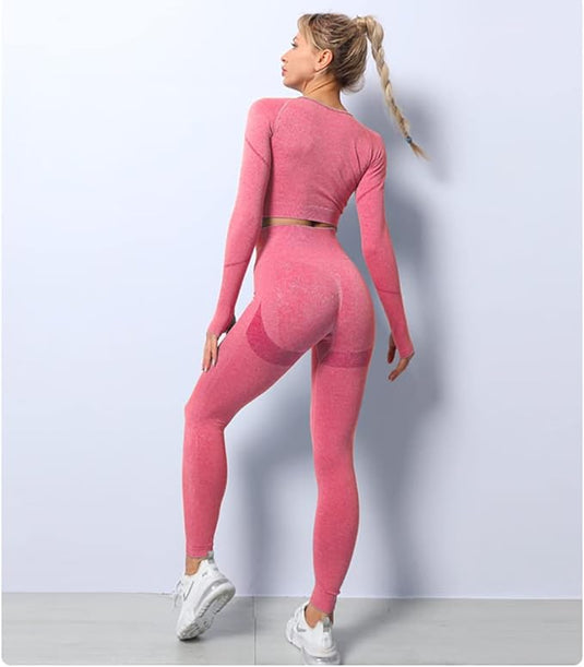 Een persoon met lang gevlochten haar staat met zijn gezicht naar een muur en draagt de Zie er geweldig uit en voel je geweldig in deze 3-delige yoga workoutset!, die een roze crop top en een karakteristieke legging bevat. Ze staan op een lichtgrijze vloer, sportieve witte sneakers gemaakt van geweven stof voor comfort.