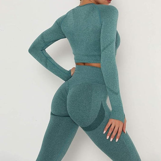 Een persoon in een blauwgroene outfit met lange mouwen uit de 3-delige yoga workoutset, gemaakt van slijtvaste stof, staat bij een witte muur met één hand op de heup, en legt moeiteloze elegantie vast.