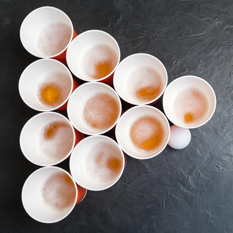 Laad afbeelding in Galerijviewer, Een driehoekige opstelling van gedeeltelijk gevulde rode kopjes met een witte bal op een donkere ondergrond benadrukt de essentie van de Speel, dag uit en knutsel met deze handige set van 100 bierpongballetjes in verschillende drankspelletjes.

