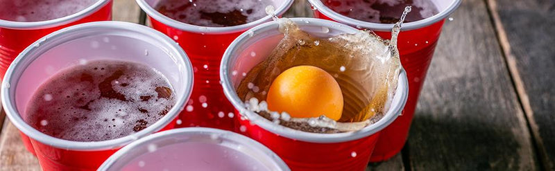 Laad afbeelding in Galerijviewer, Een gele bal uit de &quot;Speel, dag uit en knutsel met deze uitgebreide set van 100 bierpongballetjes!&quot; spat in een met rood plastic bier gevulde beker. Andere soortgelijke kopjes worden vlakbij op een houten oppervlak geplaatst.
