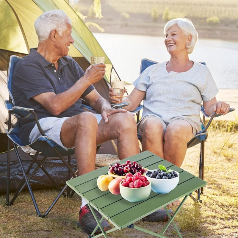 Load image into Gallery viewer, Een ouder echtpaar zit in kampeerstoelen en deelt een toost met een drankje. Voor hen staat onze &quot;Ontdek de ultieme kampeerervaring met onze draagbare campingtafel!&quot;, beladen met schalen vol kersen, bessen en perziken. Een tent en waterpartij op de achtergrond versterken de rust en charme van het tafereel.
