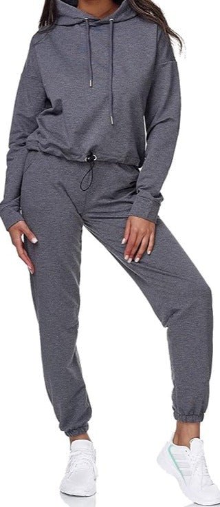 Laad afbeelding in Galerijviewer, Een persoon in een grijs &quot;Dames joggingpak: Comfort en stijl in één&quot; staat met één hand in een zak en de andere op de heup, wat de comfortabele pasvorm benadrukt. Witte sneakers gemaakt van hoogwaardige materialen maken de stijlvolle, relaxte look compleet.
