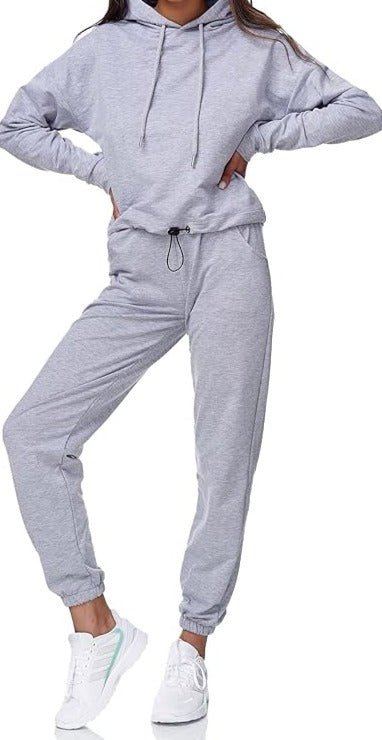 Met de Dames joggingpak: Comfort en stijl in één, staat een persoon zelfverzekerd met één been gebogen, gehuld in een grijze hoodie en bijpassende joggingbroek voor een comfortabele pasvorm, gecombineerd met kraakwitte sneakers.