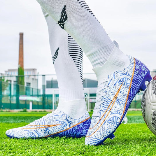 Een persoon in paars-witte "Domineer het veld" voetbalschoenen met uitstekende grip en stabiliteit staat op een grasveld naast een voetbal, met witte sokken met zwarte designs. Op de achtergrond zijn een hoge schoorsteen en een sportveld te zien.