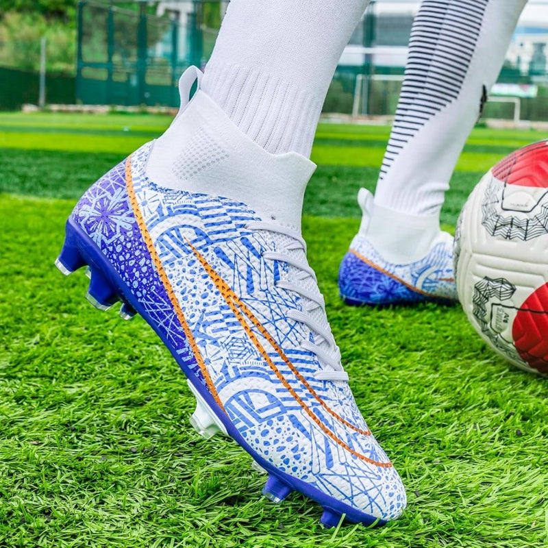Laad afbeelding in Galerijviewer, Close-up van een persoon die hoogwaardige voetbalschoenen draagt op een grasveld. Het witte ontwerp met blauwe en oranje patronen en de hoge schoenen voor grip en stabiliteit zijn goed te zien, naast een rood-witte voetbal.
