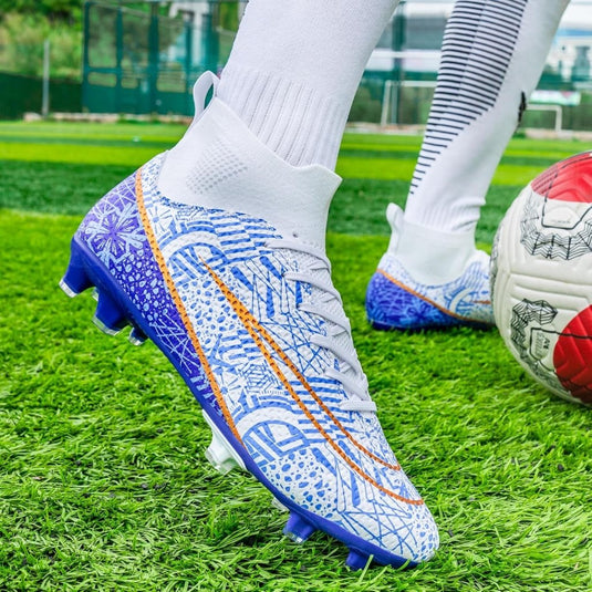 Close-up van een persoon die hoogwaardige voetbalschoenen draagt op een grasveld. Het witte ontwerp met blauwe en oranje patronen en de hoge schoenen voor grip en stabiliteit zijn goed te zien, naast een rood-witte voetbal.