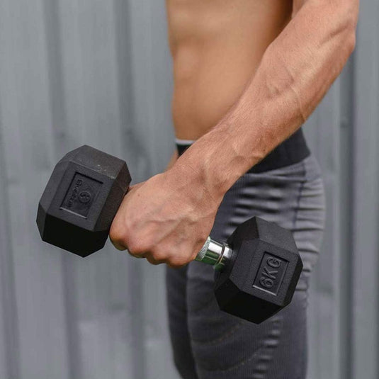 Een persoon pakt de Ultieme hexagonale 6 kg halter vast, geprezen om zijn ergonomische ontwerp en duurzame rubber, staande tegen een grijze houten achtergrond.