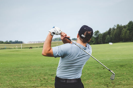 Een persoon draagt een blauw shirt en een pet en speelt golf. Hij zwaait met een club op een grasveld met bomen op de achtergrond.