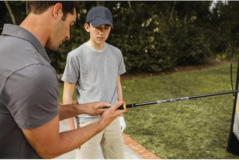 Laad afbeelding in Galerijviewer, Een volwassen golfer gebruikt de Perfecte grip met de golfvliegtrainer om een techniek te demonstreren aan een jongere met een pet en een gestreept shirt, wat mogelijk een inspiratie kan zijn voor de volgende generatie in een buitenomgeving.
