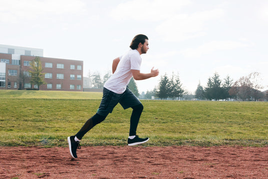 Een persoon rent over een baan in sportkleding, waaronder een wit shirt, een zwarte korte broek en een legging, met een grasveld en een gebouw op de achtergrond.