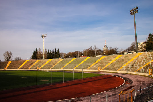 Een leeg openluchtstadion met een atletiekbaan, groen veld en tribunes onder een helderblauwe lucht. Schijnwerpers zijn aanwezig langs de omtrek.