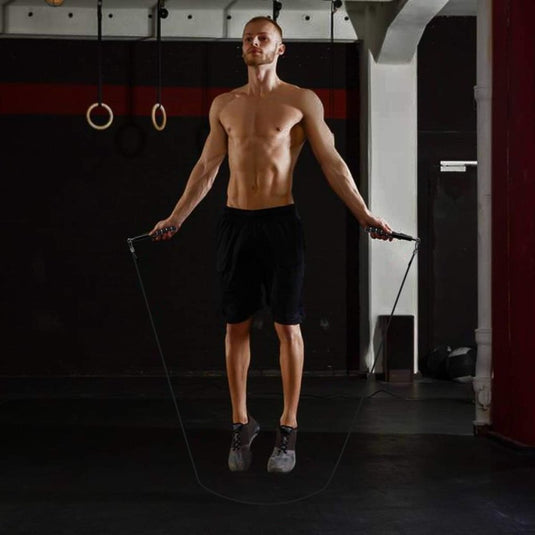 Een shirtloze man in een zwarte korte broek springt binnenshuis touw te midden van gymspullen zoals ringen, waarbij hij vakkundig gebruik maakt van het "Maximaliseer je training met het verstelbare springtouw", dat is voorzien van een antislip aluminium handvat voor veilige grip en nauwkeurige trainingsbewegingen.