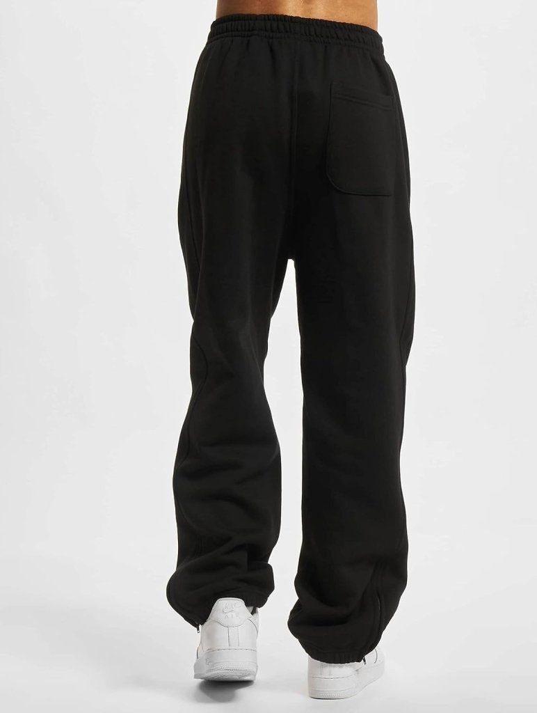 Laad afbeelding in Galerijviewer, Een persoon wordt van achteren getoond met de &quot;Comfortabele en stijlvolle sweatpants voor heren,&quot; met een baggy zwarte stijl, een elastische tailleband, een achterzak en een nonchalante slanke snit. Ze worden gecombineerd met witte sneakers.
