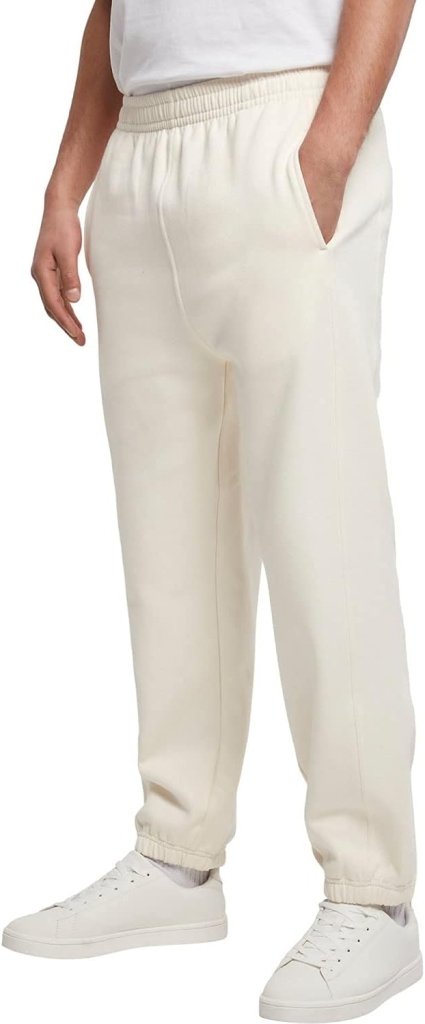 Een persoon in een comfortabele, stijlvolle, gebroken witte joggingbroek voor heren met een relaxte, strakke pasvorm en witte sneakers, staat met één hand in de zak tegen een effen achtergrond.