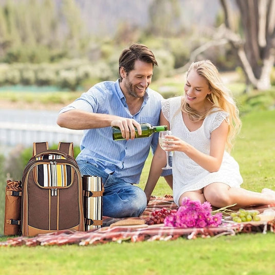 Een man en een vrouw zitten op een picknickkleed bij het serene meer terwijl hij haar een drankje inschenkt. Naast hen staat de Picknickrugzak voor 4 personen voor het ultieme picknickplezier, gevuld met premium picknickbenodigdheden, die een elegante touch toevoegt aan hun outdoor-uitje.