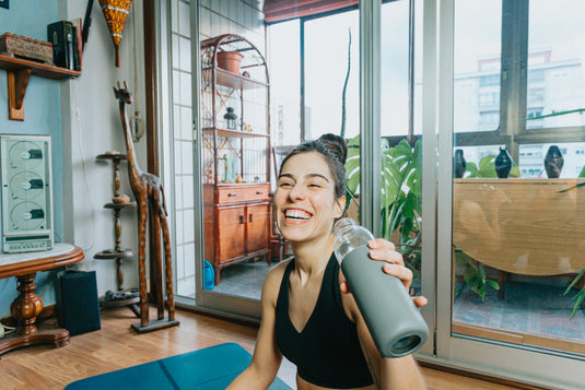 Een vrouw in sportkleding lacht terwijl ze binnen een fles water vasthoudt, met uitzicht op een balkon en planten op de achtergrond.
