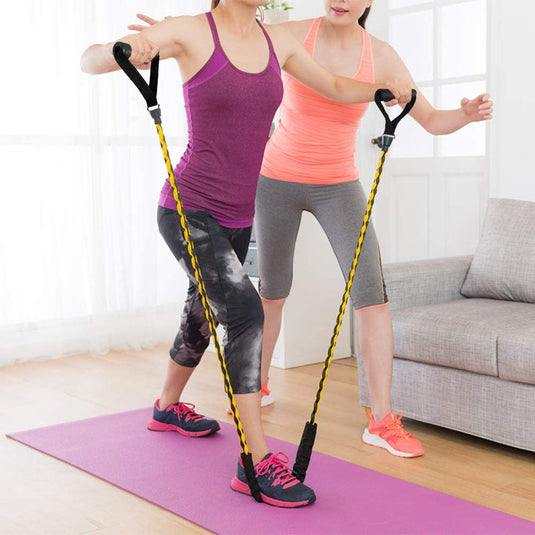 Twee vrouwen oefenen met weerstandsbanden op een paarse mat in een woonkamer. De een doet lunges naar voren terwijl de ander assisteert, waarbij ze zich concentreren op het vergroten van spierkracht. Ze dragen sportkleding en genieten van de comfortabele grip van de "Ontdek de ultieme weerstandsbanden voor maximale kracht en comfort.