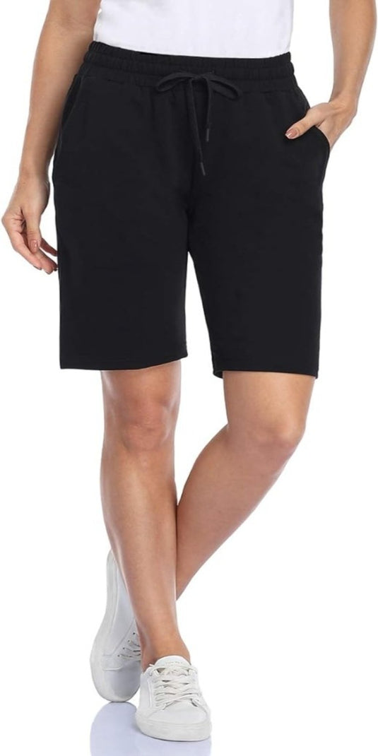 Een persoon in een wit overhemd en een "Bermudashort voor dames: comfortabel, veelzijdig en perfect voor elke gelegenheid" staat met gekruiste benen, handen in de zakken, gecombineerd met witte sneakers voor een casual look.