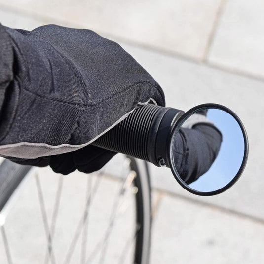 Een gehandschoende hand houdt het fietsstuur vast, waarbij een Verbeter je fietstocht met de brede BM-45 stuureinde fietsspiegel de weg erachter weerspiegelt - een perfecte mix van duurzaamheid en functionaliteit.