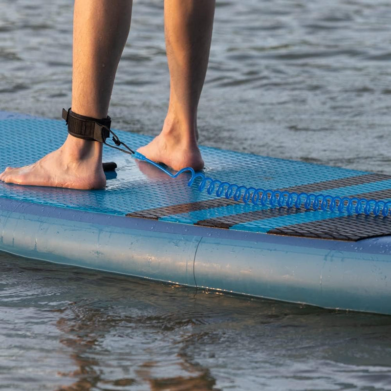 Laad afbeelding in Galerijviewer, Een persoon staat zelfverzekerd op een blauw paddleboard, met de Coiled SUP leash: de veiligste manier om te genieten van het water, stevig om de enkel gewikkeld voor de veiligheid.
