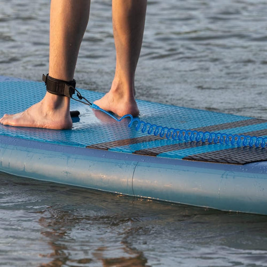 Een persoon staat zelfverzekerd op een blauw paddleboard, met de Coiled SUP leash: de veiligste manier om te genieten van het water, stevig om de enkel gewikkeld voor de veiligheid.