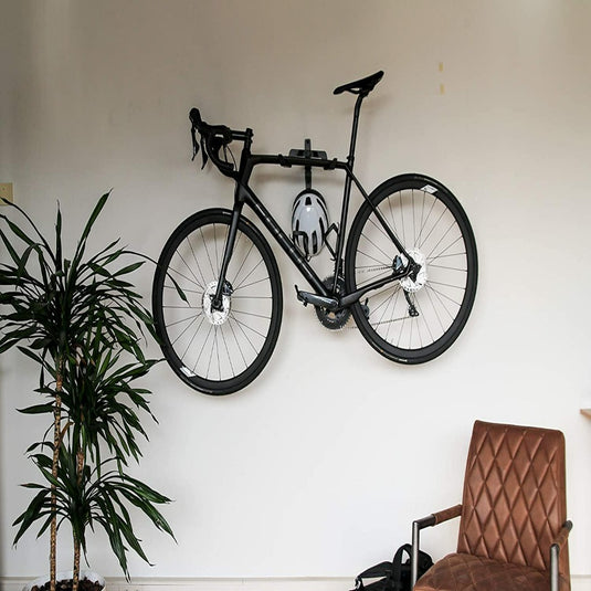 Een fiets is aan een witte muur bevestigd met behulp van een "Optimaliseer je ruimte met onze stijlvolle fiets ophangsysteem," met een grijze helm eronder. Links staat een plant en rechts staat een bruine gewatteerde stoel.