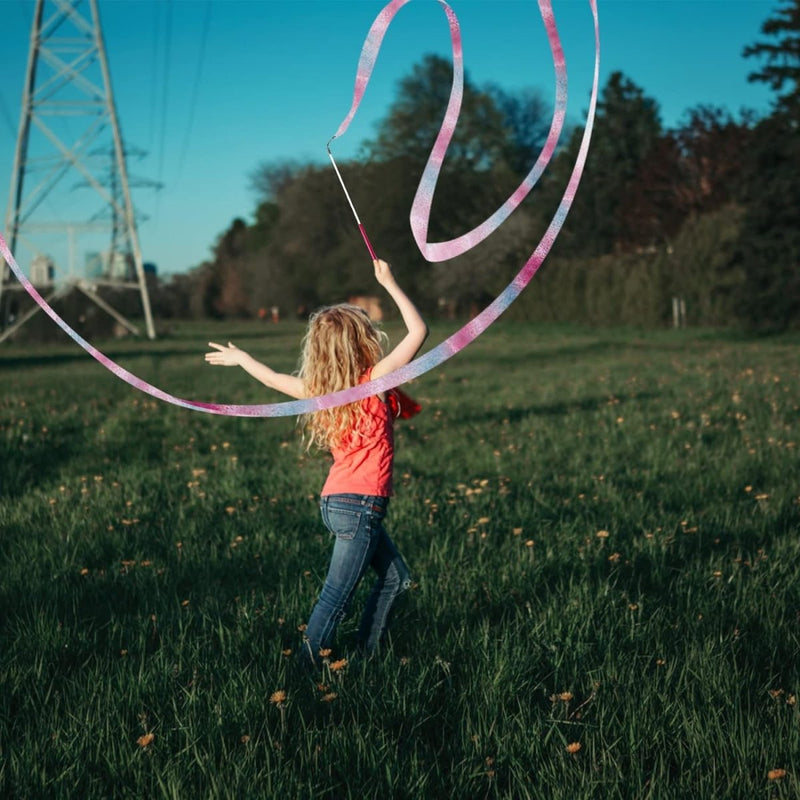 Load image into Gallery viewer, Een kind met lang haar draagt een rood shirt en draait sierlijk een roze lint in een grasveld met bomen en verre elektriciteitsleidingen, met behulp van Dansbanden voor kinderen: De ultieme manier om je dansroutines naar een hoger niveau te tillen.

