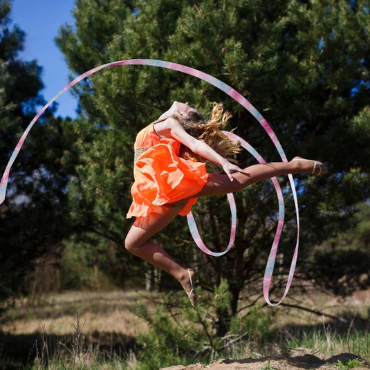 In een hemelse gratie voert een danseres in een lichte oranje polyester jurk met Dansbanden voor kinderen een adembenemende sprong uit tegen een serene achtergrond van bomen en gras, die doet denken aan ritmische gymnastiek.