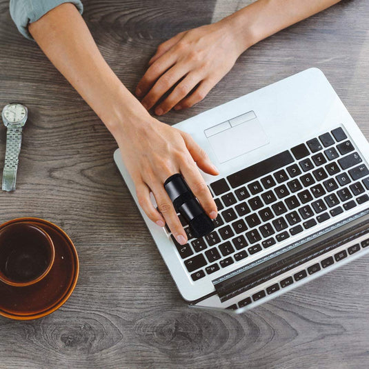 Met behulp van een draadloze ringmuis op een laptop boven een houten tafel manoeuvreert iemand behendig, ondanks het dragen van de Beste vinger spalk voor herstel en verlichting, met een horloge en koffie in de buurt.