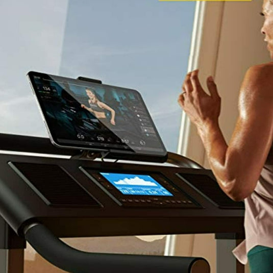 Een persoon gebruikt de Loopband en hometrainer tablethouder voor een rustige work-out, waarbij hij of zij op een loopband rent terwijl hij of zij de handen vrij heeft, terwijl hij of zij een video bekijkt op een aangesloten tablet, terwijl het zonlicht door het raam naar binnen schijnt.