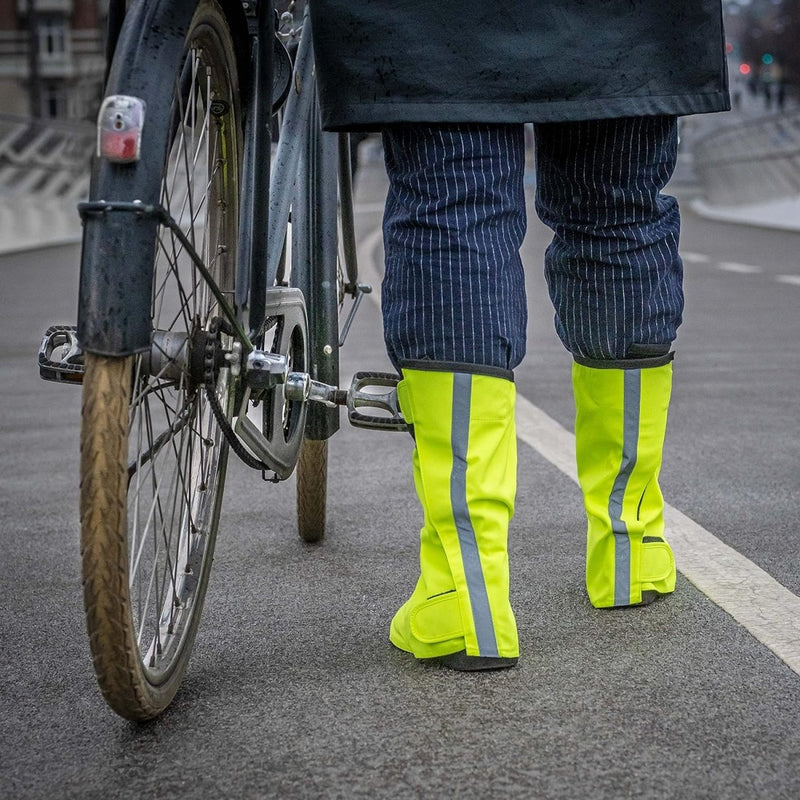 Laad afbeelding in Galerijviewer, Een persoon staat zelfverzekerd naast een fiets op de verharde weg, met felgele reflecterende regenoverschoenen voor fietsers: blijf droog en stijlvol, die veiligheid en bescherming belichamen.
