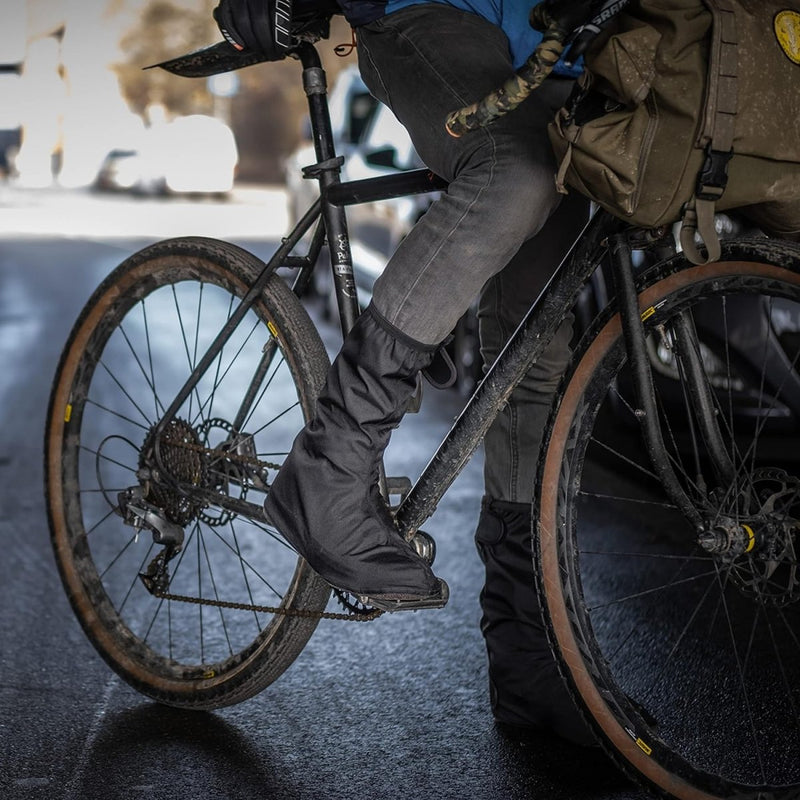 Laad afbeelding in Galerijviewer, Een persoon op een zwarte fiets trapt zelfverzekerd in regenoverschoenen voor fietsers, gekleed in een donkere spijkerbroek. Het blauwe jack biedt extra bescherming en een groene tas is stevig aan de fiets bevestigd.
