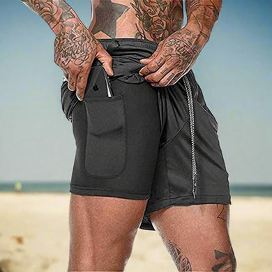 Een getatoeëerde persoon staat op het strand en draagt onze ultieme comfortabele 2-in-1 shorts voor heren, gemaakt van sneldrogend materiaal. Hij schuift een smartphone in zijn zak.