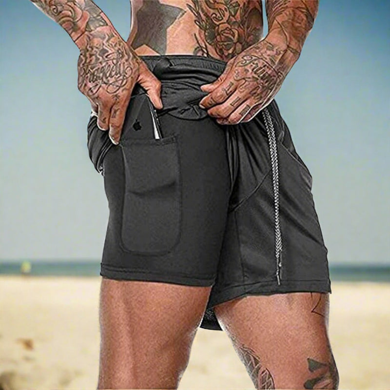 Laad afbeelding in Galerijviewer, Een getatoeëerde persoon staat op het strand en draagt onze ultieme comfortabele 2-in-1 shorts voor heren, gemaakt van sneldrogend materiaal. Hij schuift een smartphone in zijn zak.
