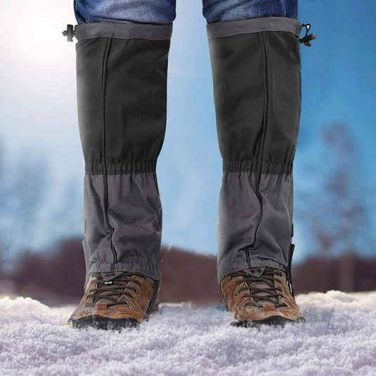 Een persoon in bruine wandelschoenen en onze volwassen sneeuwgamaschen staan op de sneeuw, perfect winddicht, met een wazig sneeuwlandschap dat een magische winterse charme toevoegt.