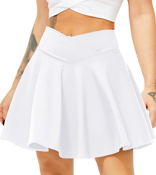 Een persoon draagt de Tennisrok met broek, een witte uitlopende rok met een kruisband en bijpassende crop top. Tatoeages sieren hun armen, terwijl de outfit de charme van een tennisrok combineert met ingebouwde shorts, waardoor stijl en functie moeiteloos samensmelten.