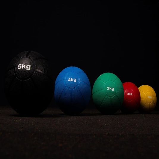 Vijf "Train je lichaam met de medicijnbal"-ballen zijn op grootte gerangschikt voor krachttraining: zwart 5 kg, blauw 4 kg, groen 3 kg, rood 2 kg en geel 1 kg. Elke bal rust op een donkere ondergrond.