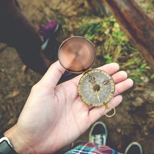 Een hand houdt de Vintage koperkompas vast: een klassiek hulpmiddel voor outdoorliefhebbers, die kardinale richtingen onthult, met het koperen deksel ernaast. Het robuuste, compacte ontwerp rust tegen een met gras begroeide achtergrond en een wazige voet.