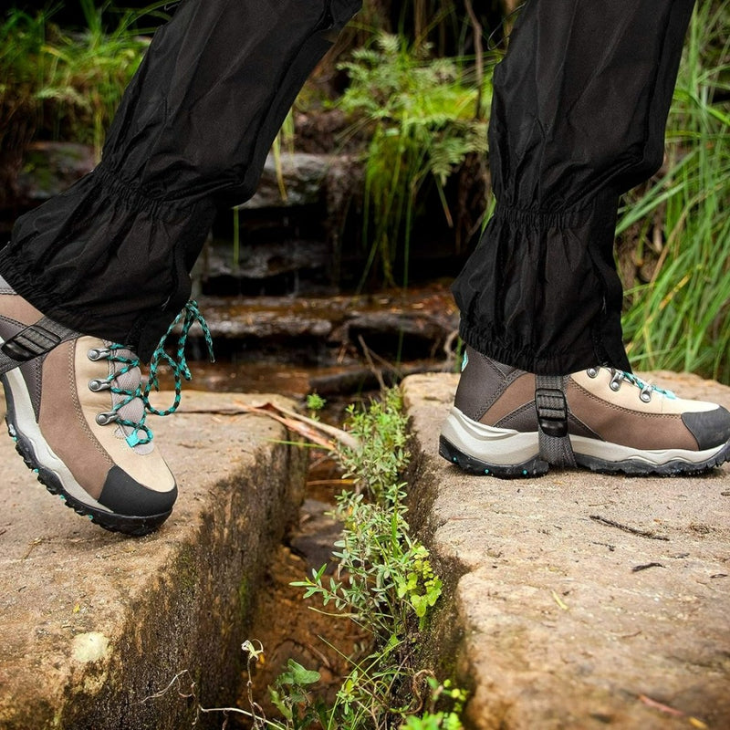 Load image into Gallery viewer, Een persoon draagt beige en zwarte wandelschoenen met blauwgroene veters en het opschrift &quot;Gaiters: de ultieme bescherming voor al je buitenactiviteiten&quot; en steekt een kleine rotsachtige kloof over, omgeven door weelderig groen.
